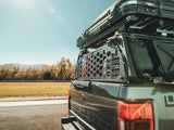 GateKEEPER — Lifestyle shot: rear three-quarter view of the installed gate with MOLLE panel accessories mounted beneath a loaded roof rack.