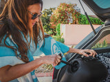 HydroPOD CARRY Portable Shower Kit — Lifestyle shot: a surfer rinses his wetsuit at his truck's tailgate, hose connected to the unit inside the bed, surfboard and boardbag nearby.