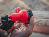 HydroPOD CARRY Portable Shower Kit — Lifestyle shot: two attendees at an overlanding expo use the hose nozzle to rinse a leashed dog beside a rooftop-tent-equipped rig.