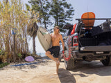 HydroPOD ACS RACK Shower Kit — Lifestyle shot: a surfer rinses sandy feet with the coiled hose stretched from the rack-mounted tank at a beach parking lot after a session.