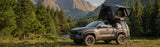 Roofnest Condor 2 Air — Lifestyle shot: open tent on a pickup at a mountain campsite in the pines, ladder down for the night