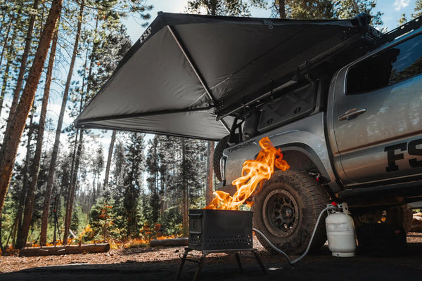 FSR 270 Awning — Lifestyle shot: deployed at a forested campsite with a propane firepit, sheltering the camp cook area