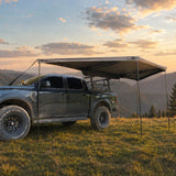 NaturNest 180° Car Awning — Lifestyle shot: awning deployed off the side of a truck at a remote overland campsite, illustrating the quick-shade setup drivers rely on between stops